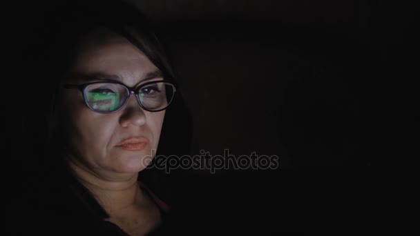 Una mujer está sentada en un sofá en una habitación oscura y trabajando en una tableta . — Vídeos de Stock