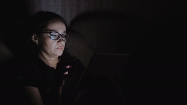 Una mujer está sentada en un sofá en una habitación oscura y trabajando en una tableta . — Vídeos de Stock