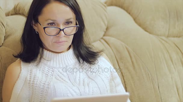 Uma mulher de óculos senta-se no sofá com um tablet nas mãos . — Vídeo de Stock