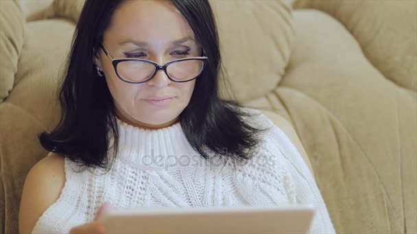 Uma mulher de óculos senta-se no sofá com um tablet nas mãos . — Vídeo de Stock
