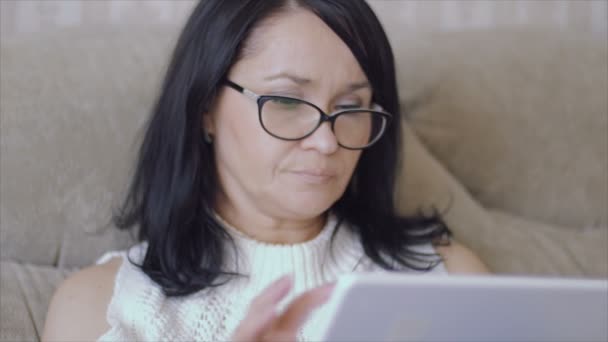 Una mujer con gafas se sienta en el sofá con una tableta en las manos . — Vídeo de stock