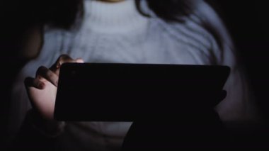 Beyaz bir ceketi giyen bir kadın onun elinde bir tablet ile karanlıkta oturmuş.