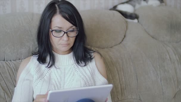 Una mujer está sentada en el sofá con una tableta en las manos . — Vídeo de stock