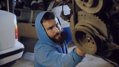 Sakallı araba tamircisi oturur ve aşağıdan zincirleri üzerinde asılı motor gövdesi içine civata vidalar.