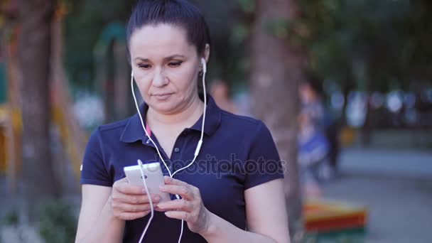 Una mujer con auriculares se para en el parque y desplaza su dedo por la pantalla del teléfono inteligente . — Vídeo de stock