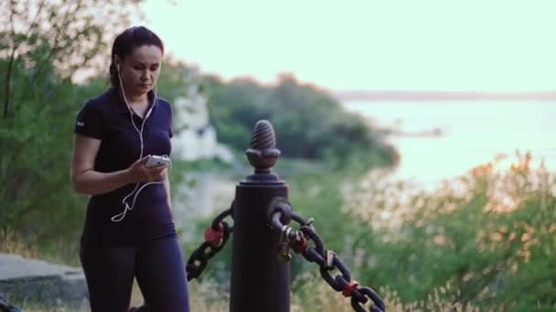 Una donna in cuffia e in uniforme sportiva cammina lungo il molo e guarda nello smartphone . — Video Stock