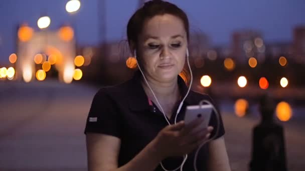 A woman stands in the evening on the embankment and scrolls the screen of her smartphone with her finger. — Stock Video