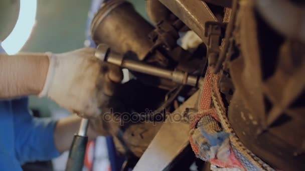 El mecánico barbudo aprieta el perno con una herramienta en el casco de un motor automotriz suspendido en un taller de reparación . — Vídeos de Stock