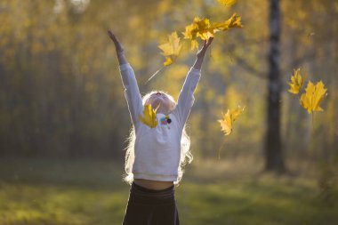 Sonbahar parkında bir kız akçaağaç yaprakları serpiyor. Sonbaharda yürüyüşe çıkan bir çocuk.