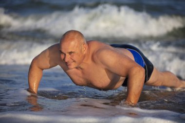 Kumsalda egzersiz yapan yaşlı bir adam. Bir adam suyun içinde yukarı itiyor. Sahilde aktif tatil. Egzersiz. Sağlıklı ve kaslı vücudu olan yakışıklı, aktif bir adamın şınav çekerken yakın çekimi. Sahilde Sportif Atletik Erkek Egzersizi, Tr.