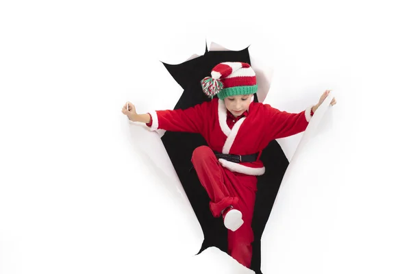 Criança Papai Noel sai de um buraco no papel. Um rapaz disfarçado de Ano Novo está a sorrir. Surpresa de férias. Conceito de Natal — Fotografia de Stock