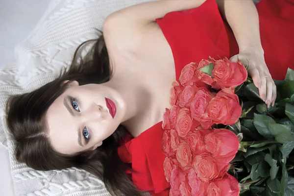 有红唇和玫瑰的漂亮模特 奢侈的女孩拿着一束花躺在床上. 穿着红衣服的年轻女人 — 图库照片