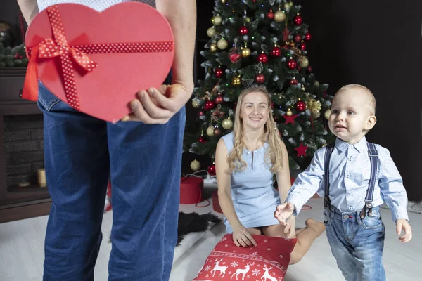 Christmas surprise. A man holds a red gift box on the background of his wife and child. Family on New Year\'s holidays. The little son runs to receive a Christmas gift.