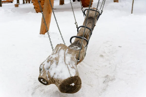 A snow-covered swing in the old style in the form of a log suspended from chains. Winter landscape.