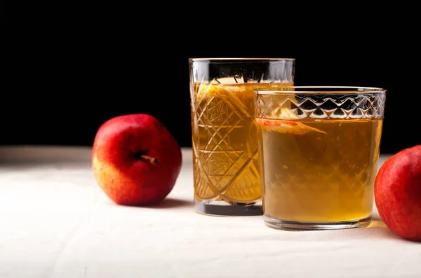 Two vintage glasses with apple cider on black background. Christmas beverages concept. Two red apples and rosemary sprig aside.  Warm backlight. Horizontal composition. Side view.