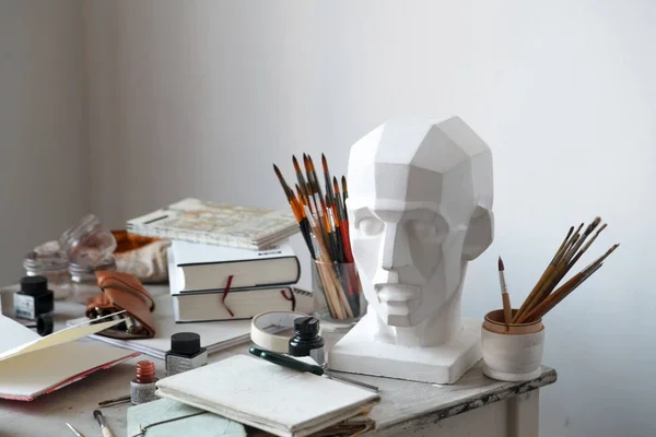 Interior of painting studio of freelance artist. Gypsum head, brushes, pencills, ink and paint bottles with sketchbooks on the table. Freelance artist lifestyle concept. Copy space