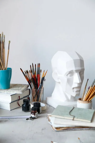 Interior of painting studio of freelance artist. Gypsum head, brushes, pencills, ink and paint bottles with sketchbooks on the table. Freelance artist lifestyle concept. Copy space