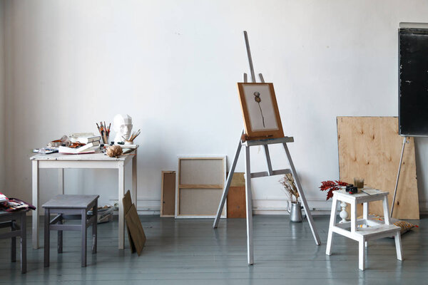 Interior of painting studio of freelance artist. Gypsum head, brushes, pencills, ink and paint bottles with sketchbooks on the table. Easel with watercolor painting. Freelance artist lifestyle concept. Copy space