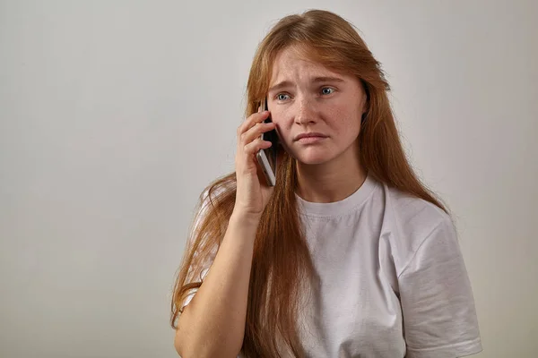 portrait of young red-headed woman with freckles holding phone next to ear and talking with sad expression on face