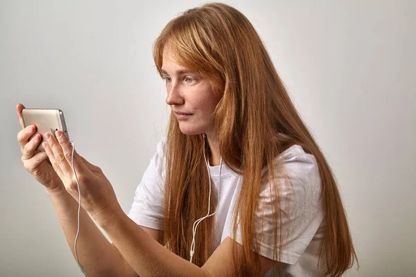 young red-headed woman with freckles holding phone in horizontal position with plugged in earphones
