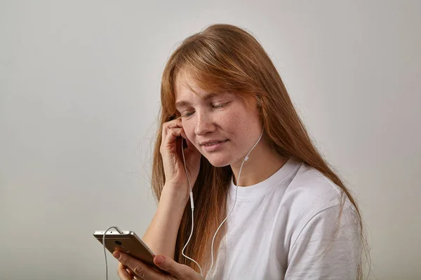 young red-headed woman with freckles holding phone while answering video call with plugged in earphones