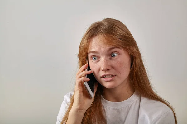 young red-headed woman with freckles holding phone next to ear and talking with surprised expression on face