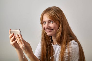 Kızıl saçlı olan kadın içinde takılı kulaklık ile yatay pozisyonda telefon tutarak ve kameraya bakarken gülümseyen çiller 