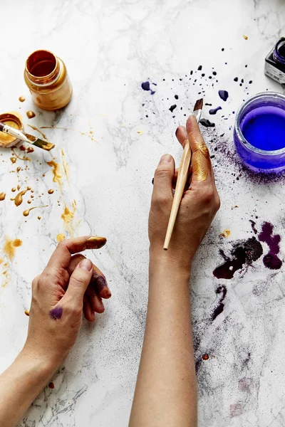 view of female artist hands soiled with golden and violet paints on white background, artist workspace concept