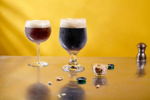 ale and stout beer in glasses with peanuts in glass bowl on metallic and yellow background