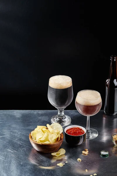 ale and stout beer in glasses with sauce and chips in bowls on metallic and black background