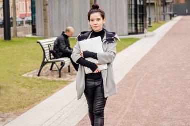 Asyalı kadın dizüstü elinde tutan ve şehir parkında yürüyüş gri palto giyiyor 