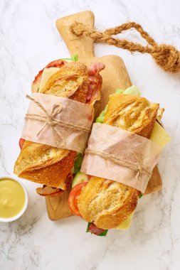 Pastırmalı, peynirli, sebzeli, ahşap kesim tahtasıyla mermer arka planda salatalı taze baget sandviçler.