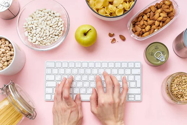 Creative layout with pantry contents and female hands typing on keyboard prepared to survive Coronavirus COVID 19 pandemic at time of cuarentine and home isolation