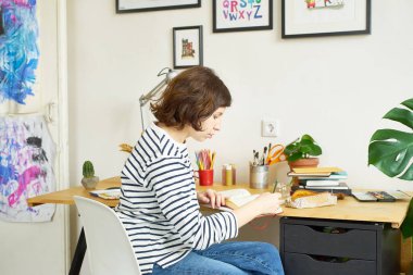 İş yerindeki kadın sanatçı evden çalışıyor. Kot pantolon ve çizgili gömlek giymiş genç bir kadın masada oturuyor ve bir illüstrasyon yaratıyor.. 