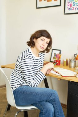 İş yerindeki kadın sanatçı evden çalışıyor. Kot pantolon ve çizgili gömlek giymiş bir kadın masada oturuyor ve resim çiziyor.. 