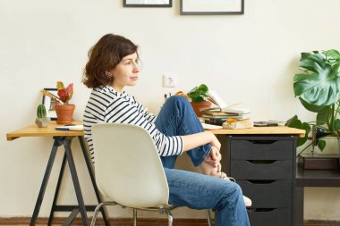 İş yerindeki kadın sanatçı evden çalışıyor. Kot pantolon ve çizgili gömlek giymiş bir kadın masada oturuyor ve resim çiziyor. İlham konsepti arıyor.