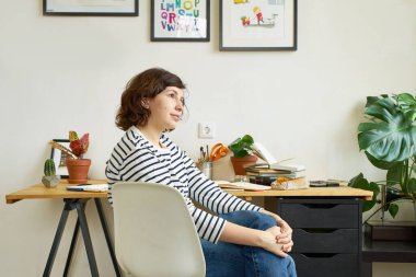 İş yerindeki kadın sanatçı evden çalışıyor. Kot pantolon ve çizgili gömlek giymiş bir kadın masada oturuyor ve resim çiziyor. İlham konsepti arıyor.
