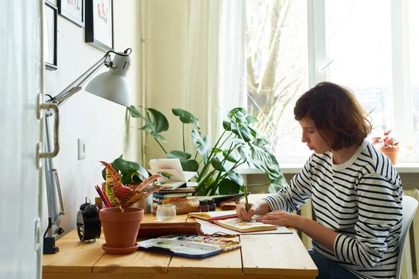 예술가 집에서 일하고 있습니다 청바지와 셔츠를 여자가 테이블에 그림을 그리는 — 스톡 사진