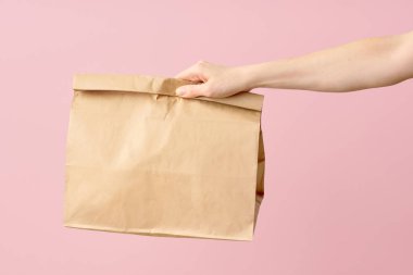 Kadın eli, pembe arka planda yiyecek teslimatı için kahverengi boş kağıt torbayı tutuyor. Paketleme şablonu fast food 'u taklit eder. 