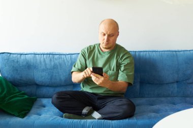 Oturma odasında mavi koltukta oturan bir adam akıllı telefon kullanıyor.