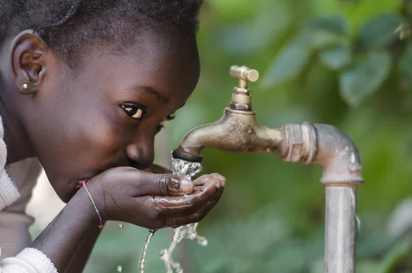 Prachtige Afrikaanse kind drinken uit een kraan (waterschaarste symbool). Afrikaanse meisje schoon drinkwater uit een kraan. Water gieten van een kraan in de straten van de Afrikaanse stad Bamako, Mali. — Stockfoto