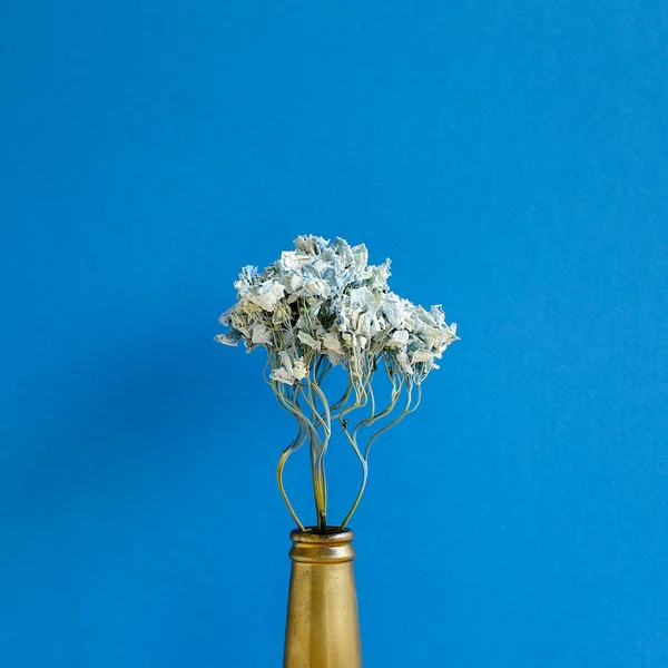 Dried blue hydrangea flower in golden bottle with blue background. Floral composition, copy space