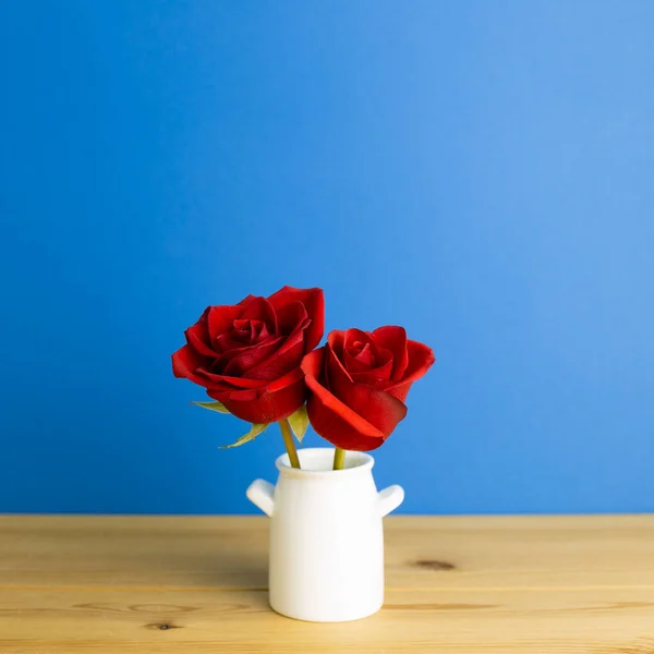 Red rose flowers in vase on wooden table with blue background. Spring floral arrangement, copy space