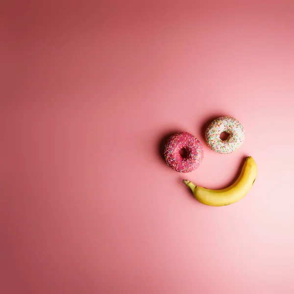 funny smiley of two donuts and banana on a pink background with place for text, an example of junk food
