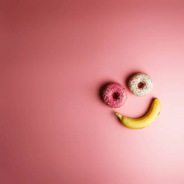 Pembe arka planda iki donut ve muzdan oluşan komik bir gülümseme. Mesaj için bir yer, abur cubur örneği.