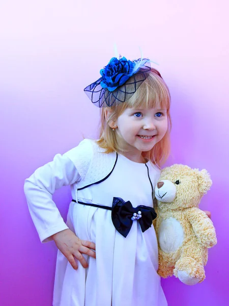 Mała dziewczynka w modny kapelusz i z zabawkami niedźwiadek w rąk, uśmiechnięty i pozowanie na tle liliowy — Zdjęcie stockowe