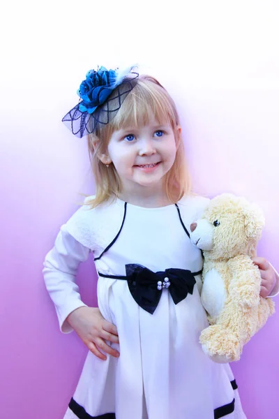 Niña en sombrero de moda y con cachorro de oso de juguete en las manos sonriendo y posando sobre fondo lila —  Fotos de Stock