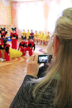 Chernihiv / Ukrayna. 28 Aralık 2018: Anaokulundaki kadın fotoğraf matinesi. Anne dijital kamerayı elinde tutuyor. Hafıza için ateş ediyorum. - Çocuk fotoğrafı. Anne kameraya bebek performansı çekiyor.