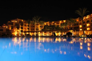 Hurghada / Egypt. 02 August 2018:Holidays at Egyptian resort. People have a rest near pool in evening. Lights of evening hotel are reflected in pool water in night. Bright lights of resort hotel. People relax in evening near pool