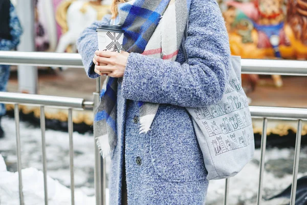 Girl in grey coat, hat and blanket scarf with cup of coffee near carousel — Stock Photo, Image